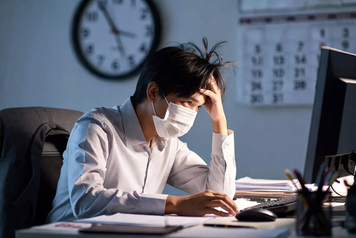 一名上班族戴著口罩在辦公桌前顯得疲憊,背景是模糊的時鐘與月曆,象徵帶病上班的壓力與時間管理